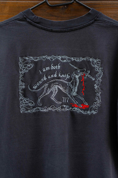 Tricou negru din bumbac organic, pe un umeraș negru, pe un fundal din lemn închis, cu o poză brodată reprezentând un lup într-o ramă decorativă, alături de textele „I am both wound and knife” și „777” brodate cu alb și detalii roșii în formă de picături de sânge.