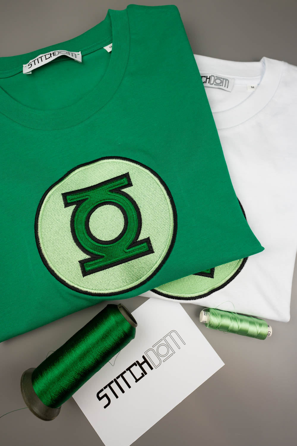 Tricou verde din bumbac organic, împăturit, cu emblema mare „Green Lantern” brodată pe piept, alături de un tricou alb, conuri de ață verde și un card cu brandul „Stitchdom”, pe un fundal gri.