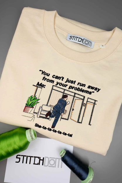 Tricou crem din bumbac organic, împăturit, cu o poză brodată reprezentând o femeie care pleacă dintr-o cameră și textul „You can't just run away from your problems”, alături de conuri de ață verde și neagră și un card alb, pe un fundal gri.