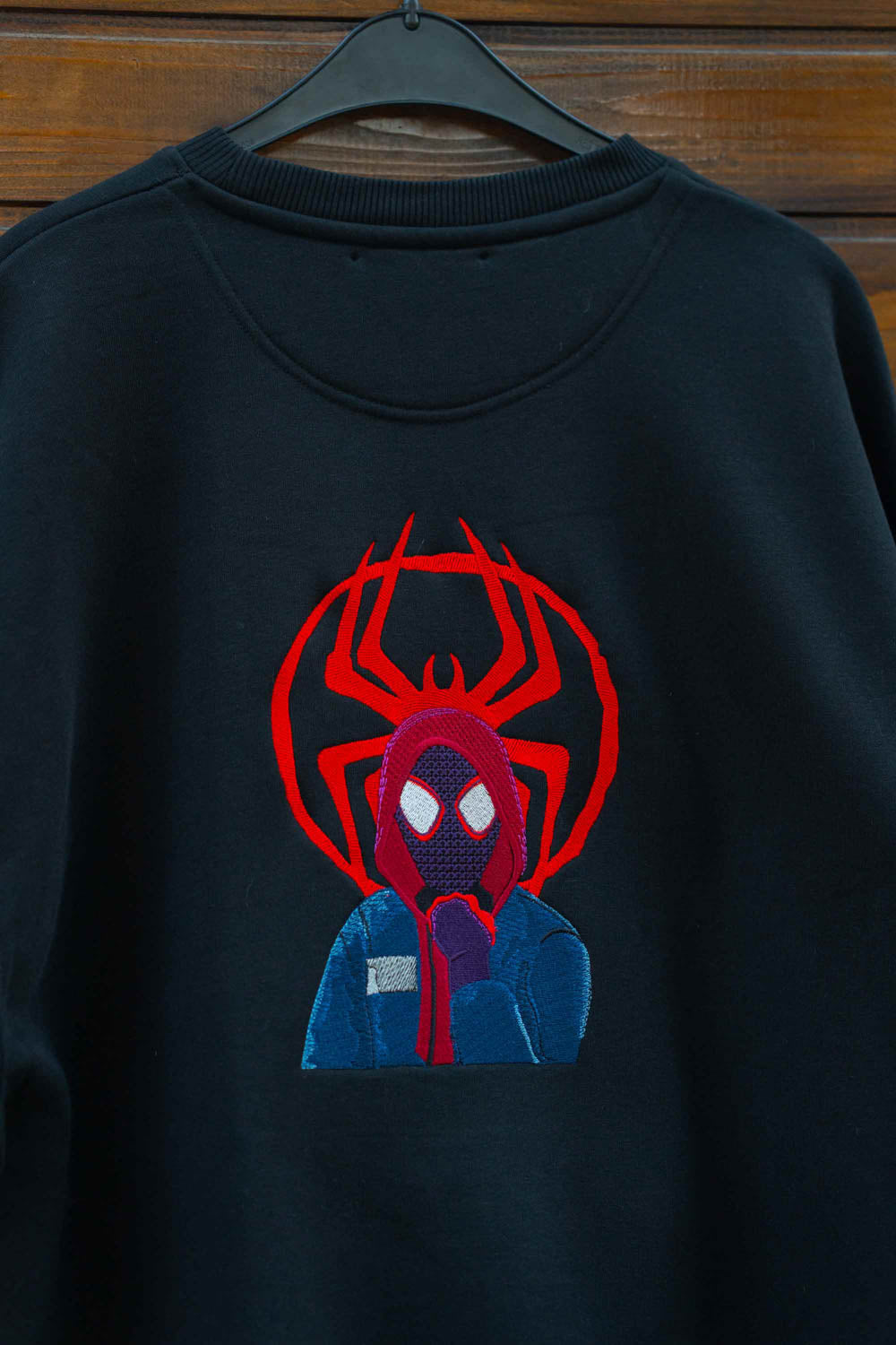 Hanorac negru din bumbac organic, cu guler rotund, agățat pe un umeraș negru, pe un fundal din lemn închis, cu un design mare brodat reprezentându-l pe Spider-Man, cu emblemă roșie de păianjen și costum albastru.