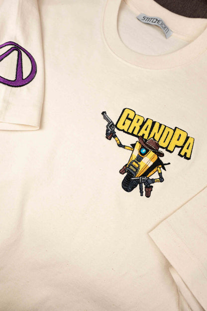Tricou alb murdar din bumbac organic, cu guler rotund, având o poză brodată reprezentând un robot cowboy galben și negru, cu textul „GRANDPA” brodat cu litere galbene îngroșate, și un semn al păcii mov brodat pe mâneca stângă, inspirat din jocul Borderlands.