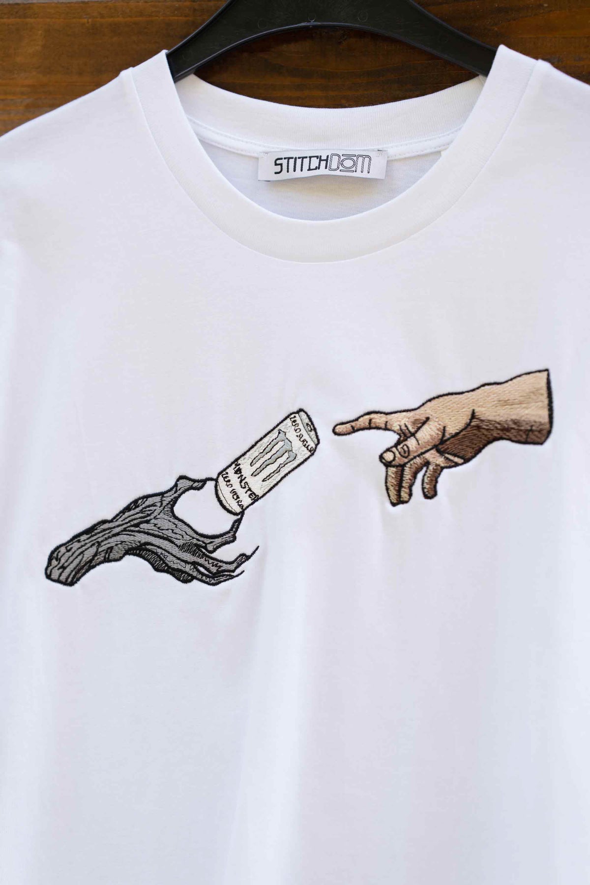 Tricou alb din bumbac organic, cu mânecă scurtă, agățat pe un umeraș negru, pe un fundal din lemn, cu o poză brodată reprezentând o mână întinsă spre o sticlă ținută de trunchiul noduros al unui copac, într-un stil inspirat de ilustrația „The Great God Marveller”.