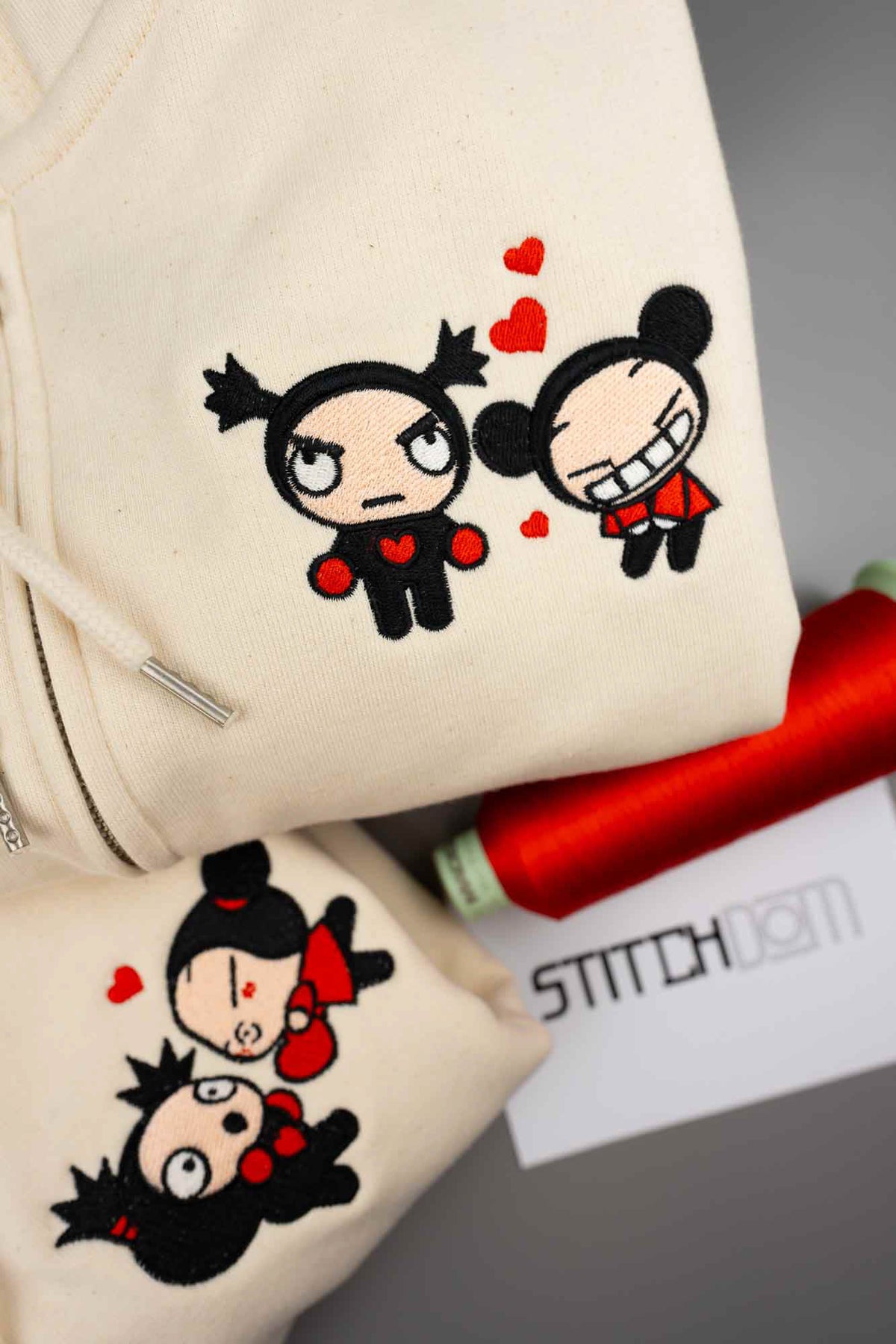 Haine crem din bumbac organic, prezentate în prim-plan, cu personaje cartoon brodate și inimioare roșii, alături de un con de ață roșie și un card cu brandul „Stitchdom”.