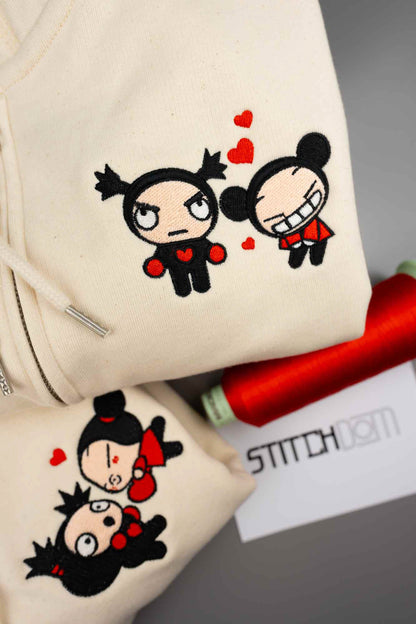 Haine crem din bumbac organic, prezentate în prim-plan, cu personaje cartoon brodate și inimioare roșii, alături de un con de ață roșie și un card cu brandul „Stitchdom”.