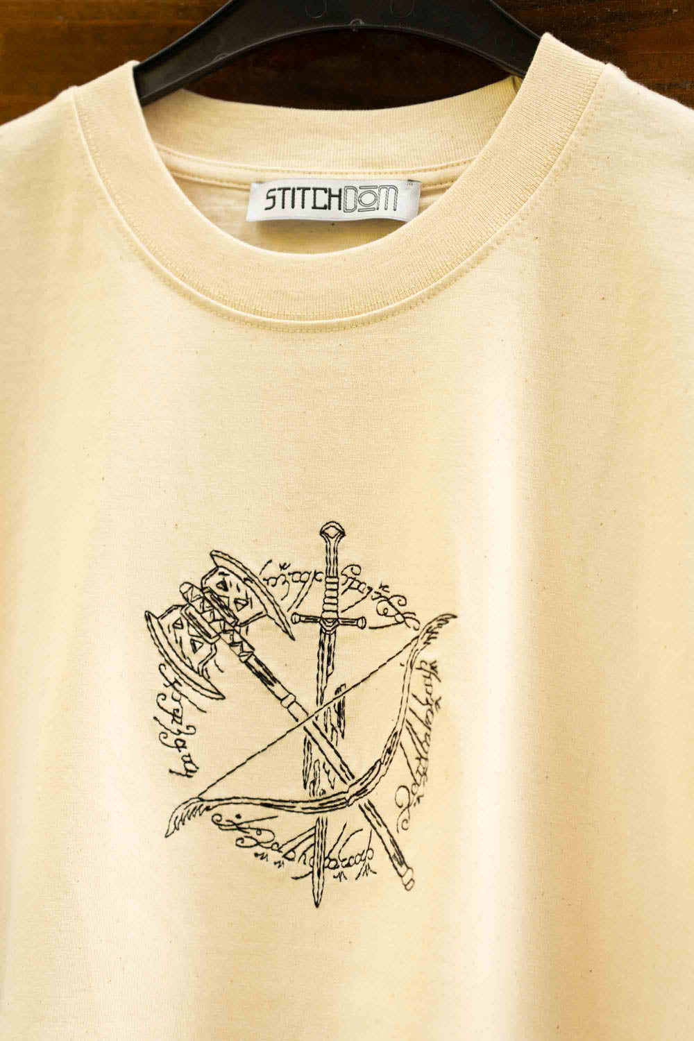 Tricou crem din bumbac organic, agățat pe un umeraș negru, pe un fundal din lemn închis, cu un design brodat negru pe piept, reprezentând săbii încrucișate, un topor de luptă și un arc, inspirat din „Lord of the Rings”, cu o etichetă albă „Stitchdom” vizibilă la gât.