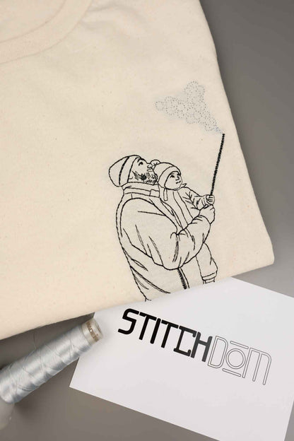 Tricou crem din bumbac organic, împăturit, cu o poză brodată tip line art, cu un părinte care ține un copil în brațe, alături de un con de ață și un card „Stitchdom”, pe un fundal gri.