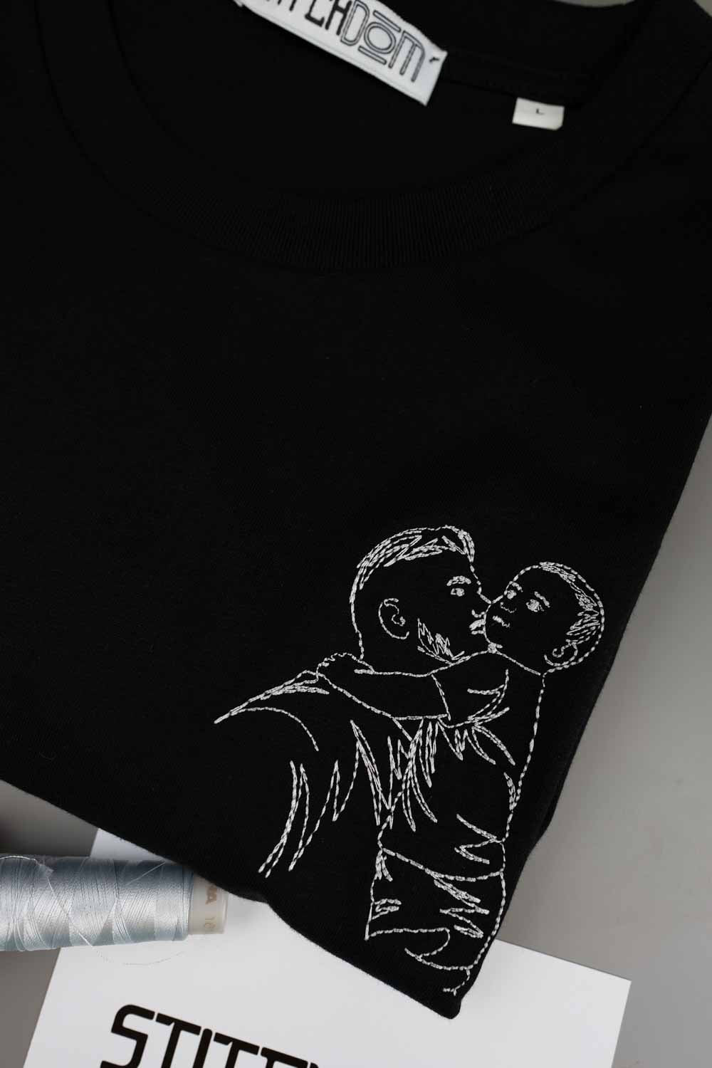 Tricou negru din bumbac organic, împăturit, cu o poză brodată tip line art, cu un părinte care ține un copil în brațe, alături de un con de ață și un card „Stitchdom”, pe un fundal gri.