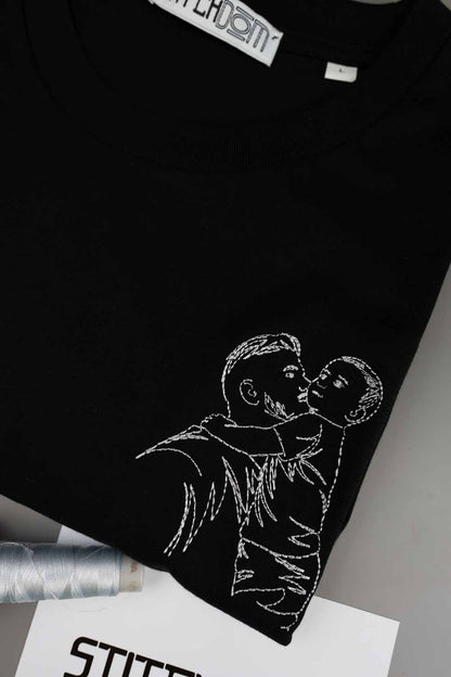 Tricou negru din bumbac organic, împăturit, cu o poză brodată tip line art, cu un părinte care ține un copil în brațe, alături de un con de ață și un card „Stitchdom”, pe un fundal gri.