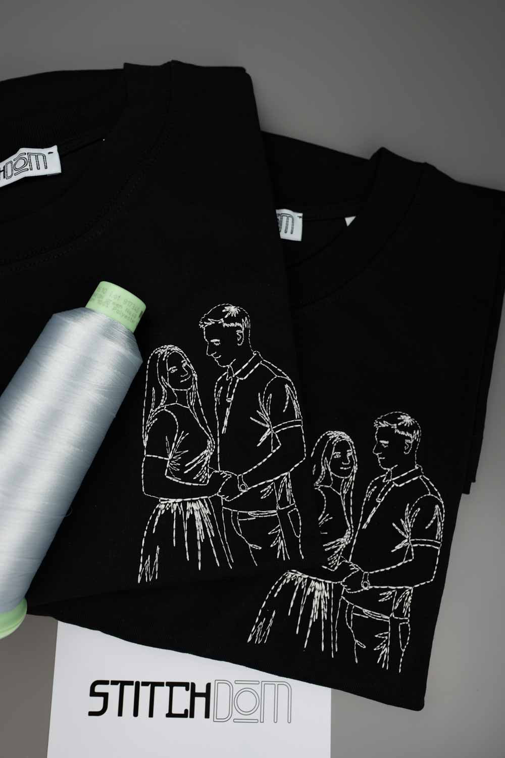 Tricou negru din bumbac organic, cu o poză brodată albă în stil line art, reprezentând un cuplu, alături de un con de ață albă și o etichetă de brand „StitchDom”, pe un fundal gri.