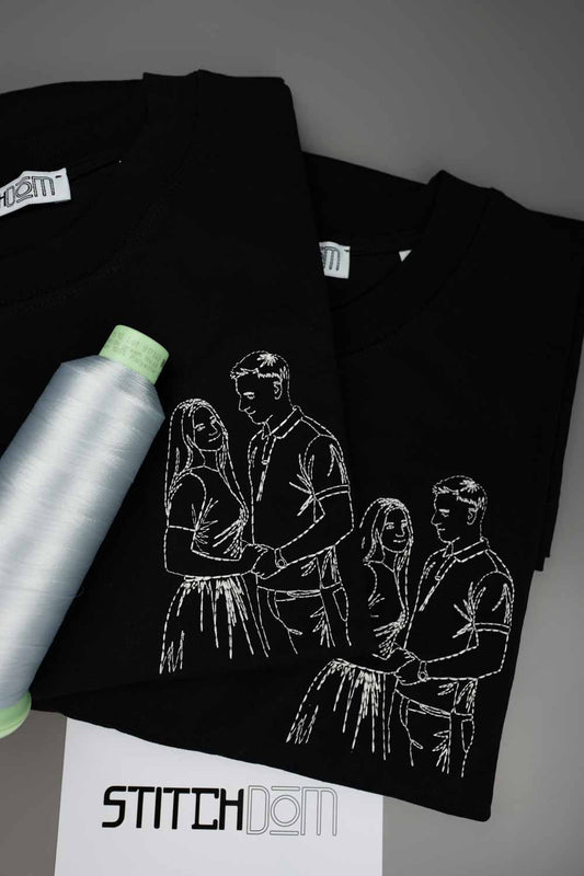 Tricou negru din bumbac organic, cu o poză brodată albă în stil line art, reprezentând un cuplu, alături de un con de ață albă și o etichetă de brand „StitchDom”, pe un fundal gri.