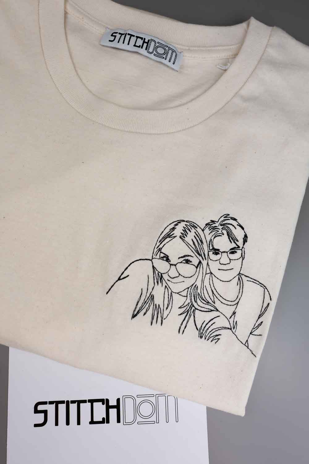 Tricou crem din bumbac organic, împăturit, cu o ilustrație neagră brodată în stil line-art, reprezentând un cuplu pe partea din față, cu eticheta interioară de la gât și un card alb pe care este vizibil logo-ul „StitchDom”.