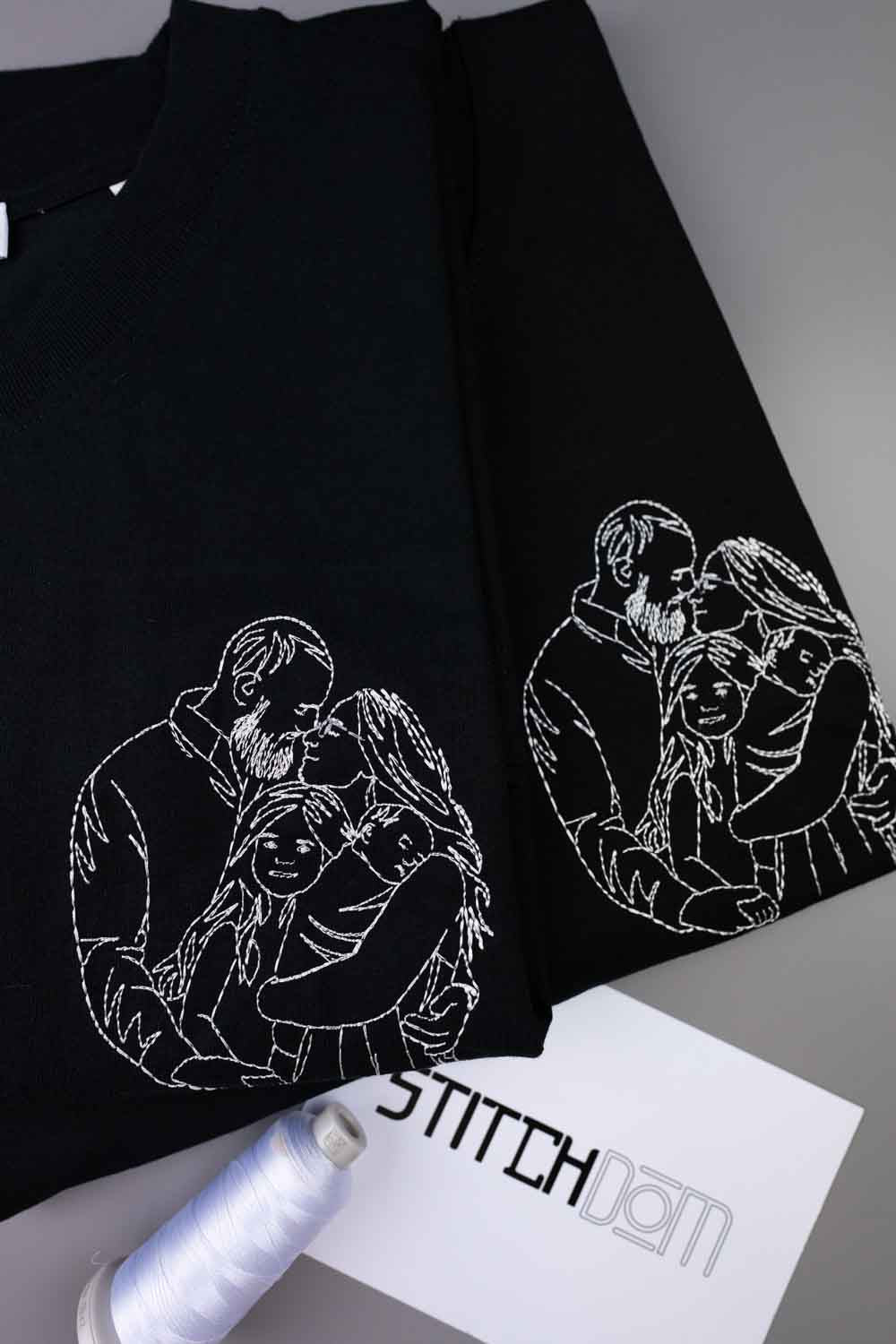 Tricou negru din bumbac organic, cu mâneci scurte, așezat pe o suprafață gri, cu o ilustrație albă brodată în stil line-art, reprezentând un cuplu și copilul lor pe piept, alături de un con de ață și un card cu brandul „STITCHDOM”, parțial vizibile în prim-plan.