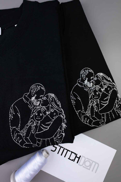 Tricou negru din bumbac organic, cu mâneci scurte, așezat pe o suprafață gri, cu o ilustrație albă brodată în stil line-art, reprezentând un cuplu și copilul lor pe piept, alături de un con de ață și un card cu brandul „STITCHDOM”, parțial vizibile în prim-plan.
