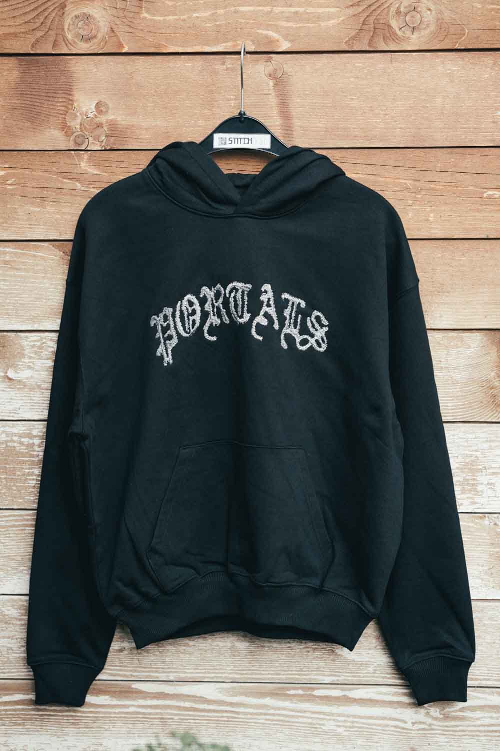 Hanorac negru din bumbac organic, pe umeraș negru, pe un fundal din lemn rustic, cu textul „PORTALS” brodat cu font gotic alb pe piept.