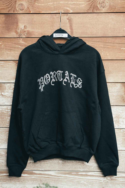 Hanorac negru din bumbac organic, pe umeraș negru, pe un fundal din lemn rustic, cu textul „PORTALS” brodat cu font gotic alb pe piept.