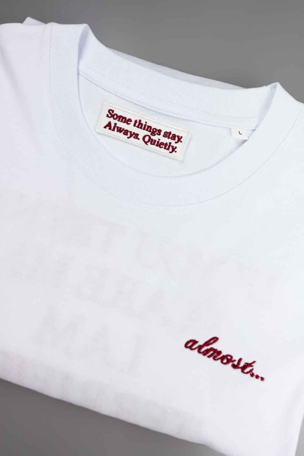 Tricou alb din bumbac organic, cu guler rotund, cu o poză discretă brodată pe piept și textul roșu „almost.” brodat.