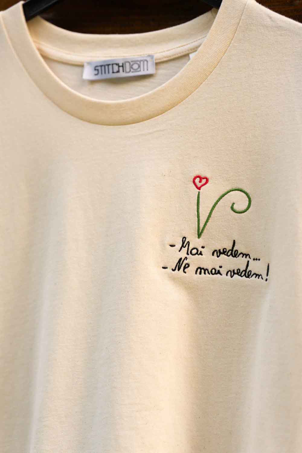 Tricou crem din bumbac organic, pe umeraș negru, cu o poză brodată pe partea stângă a pieptului, cu o tulpină verde stilizată, o floare roșie în formă de inimă și textul francez „- Mai modern.”