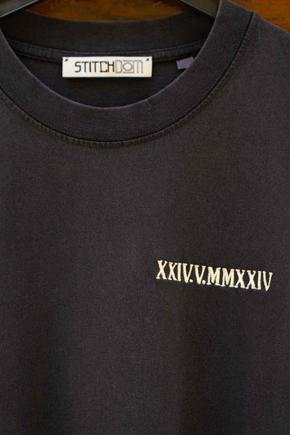 Tricou negru din bumbac organic, pe umeraș negru, cu eticheta albă „STITCHDOM” și cifre romane aurii „XXIV.V.” brodate.