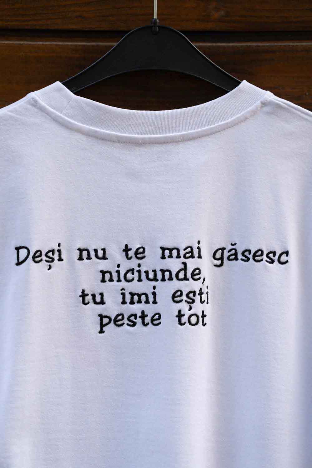 Tricou alb din bumbac organic, cu mânecă scurtă, pe umeraș negru, pe un fundal din lemn închis, cu text negru.