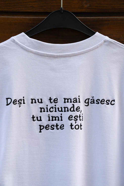Tricou alb din bumbac organic, cu mânecă scurtă, pe umeraș negru, pe un fundal din lemn închis, cu text negru.