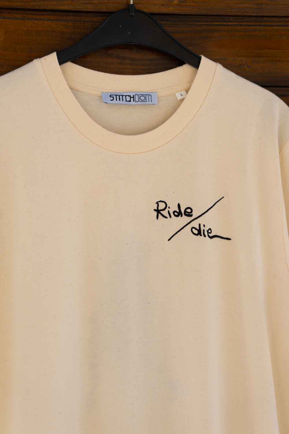 Tricou crem din bumbac organic, pe umeraș negru, pe un fundal din lemn închis, cu textul „Ride / die” brodat cu negru, în stil scris de mână, pe partea stângă a pieptului și eticheta „STITCHDOM” în interiorul gulerului.
