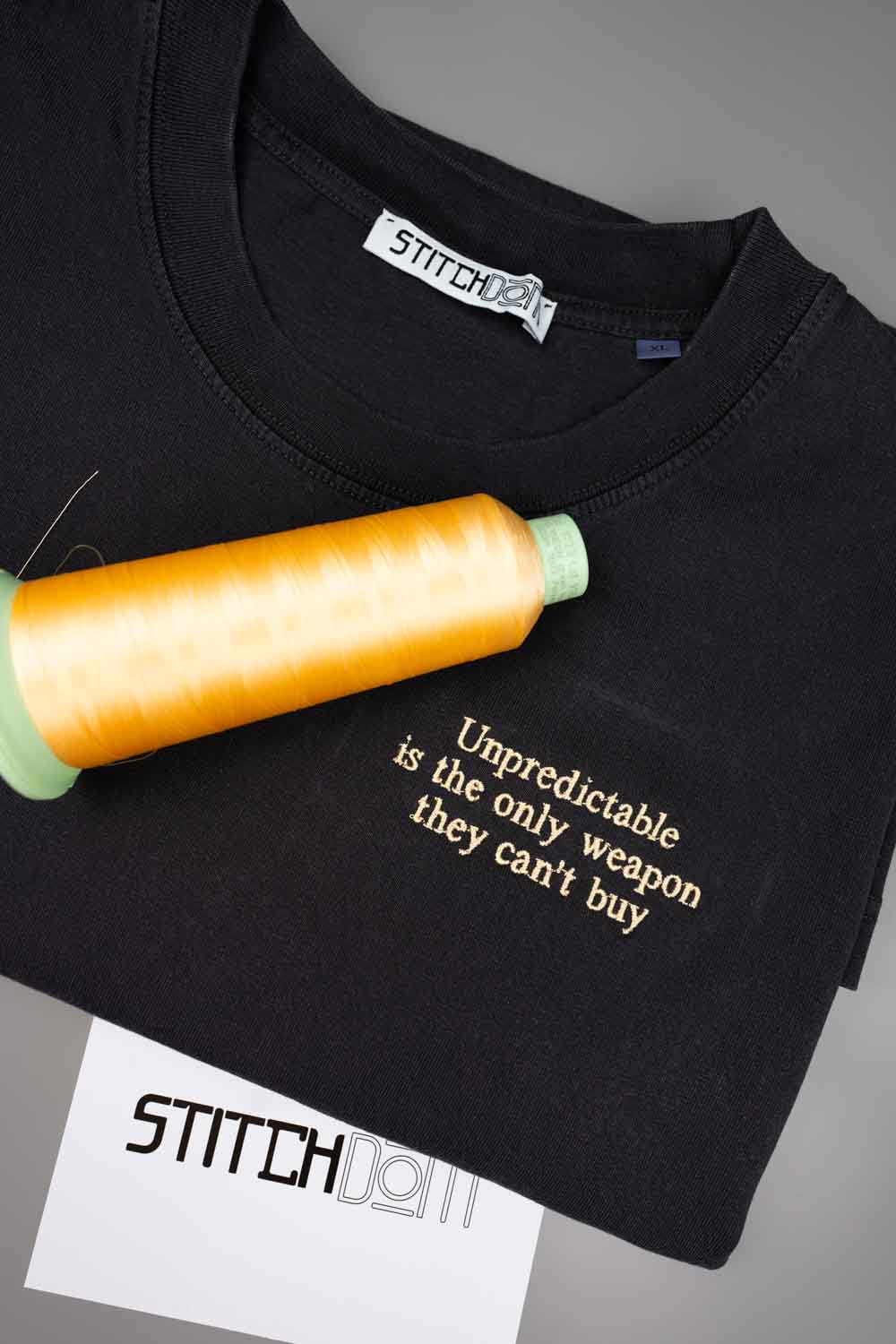 Tricou negru din bumbac organic, împăturit, cu un text amuzant brodat cu ață aurie pe piept, alături de un con de ață portocalie și un card cu brandul „StitchDOM”, pe un fundal gri.