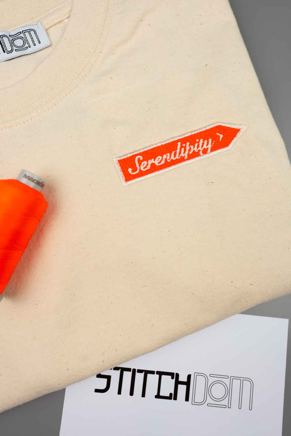 Tricou crem din bumbac organic, împăturit, cu un patch brodat portocaliu aprins pe piept, cu textul „Serendipity” brodat cu ață albă, alături de un con de ață portocalie și un card alb cu logo-ul „STITCHDOM”, pe o suprafață gri.