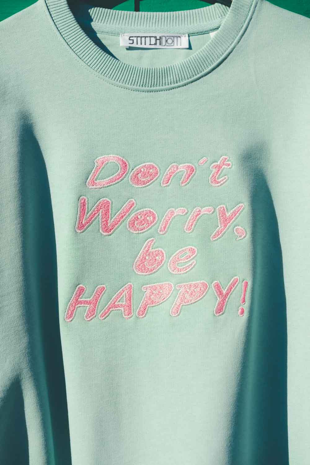 Hanorac verde mentă din bumbac organic, cu textul „Don't Worry, be HAPPY!” brodat cu roz pe piept și eticheta „STITCHDOM” vizibilă în interiorul gulerului.