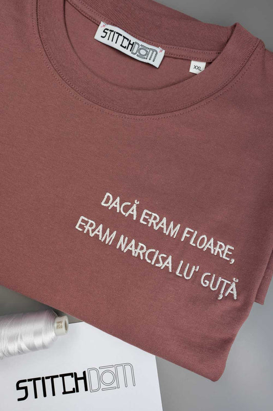Tricou roz prăfuit din bumbac organic, cu guler rotund, împăturit, cu textul alb „DACA ERAM FLOARE, ERAM NARCISA LU' GUTA” brodat pe partea din față, cu eticheta „STITCHDOM” vizibilă în interiorul gulerului, alături de un con de ață în nuanță asortată și un card cu brandul, parțial vizibile în prim-plan.