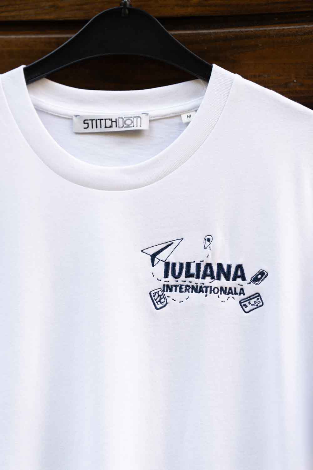 Tricou alb din bumbac organic, cu mânecă scurtă, pe umeraș negru, pe un fundal din lemn închis, cu o poză brodată bleumarin pe partea stângă a pieptului, cu un avion de hârtie și textul „IULIANA INTERNATIONALA”.