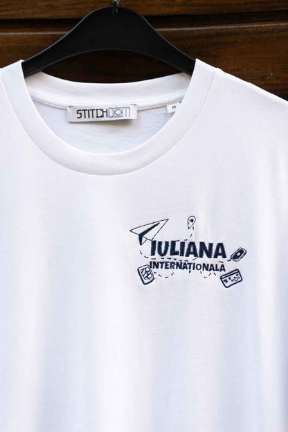 Tricou alb din bumbac organic, cu mânecă scurtă, pe umeraș negru, pe un fundal din lemn închis, cu o poză brodată bleumarin pe partea stângă a pieptului, cu un avion de hârtie și textul „IULIANA INTERNATIONALA”.