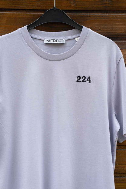 Tricou gri deschis din bumbac organic, pe umeraș negru, pe un fundal din lemn închis, cu numărul „224” brodat cu negru pe partea stângă a pieptului și eticheta „Stitchdom” vizibilă în interiorul gulerului.