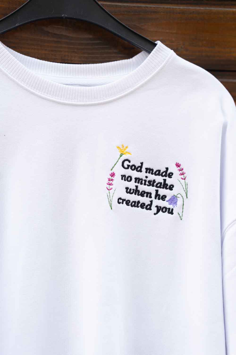 Hanorac alb din bumbac organic, pe umeraș negru, pe un fundal din lemn închis, cu o poză brodată colorată pe partea stângă a pieptului, cu textul „God made no mistake when he created you”, înconjurat de flori.