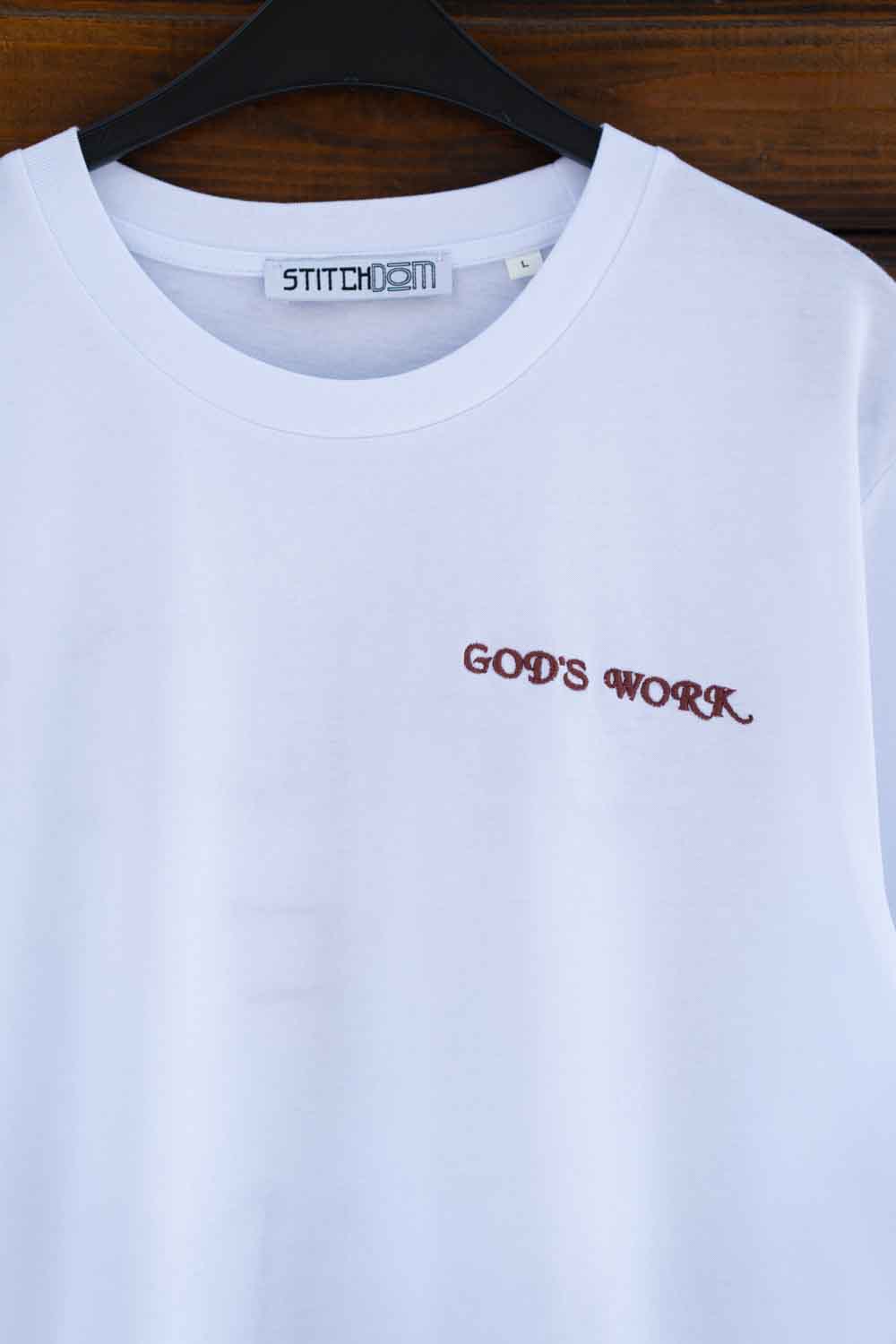 Tricou alb din bumbac organic, pe umeraș negru, pe un fundal din lemn închis, cu textul „GOD'S WORK” brodat cu roșu închis, în stil gotic, pe partea stângă a pieptului.