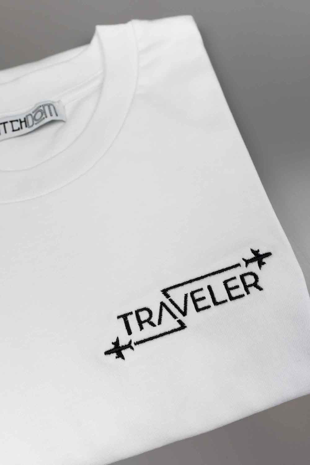 Tricou alb din bumbac organic, împăturit, pe un fundal gri neutru, cu textul „TRAVELER” brodat cu negru și simboluri cu avioane.