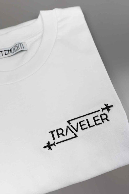 Tricou alb din bumbac organic, împăturit, pe un fundal gri neutru, cu textul „TRAVELER” brodat cu negru și simboluri cu avioane.