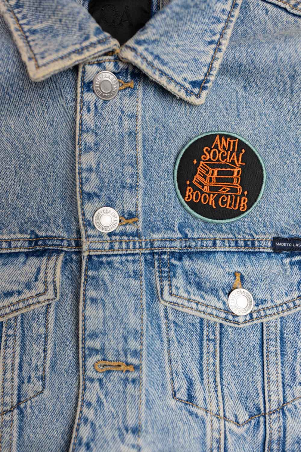 Patch brodat negru cu mesajul „Anti Social Book Club” și ilustrație de cărți, un accesoriu pentru pasionații de lectură.