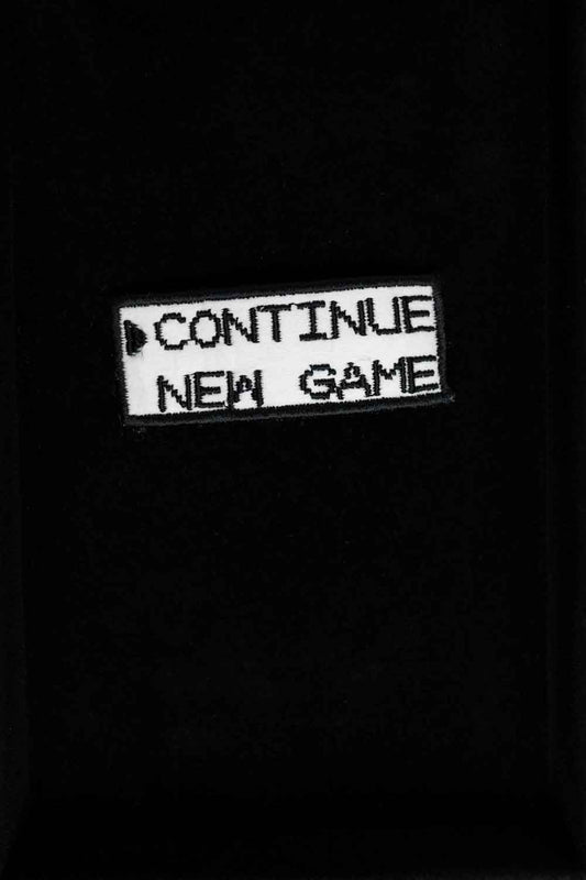 Patch brodat negru cu mesajul „Continue New Game”, un accesoriu inspirat de jocurile video retro.