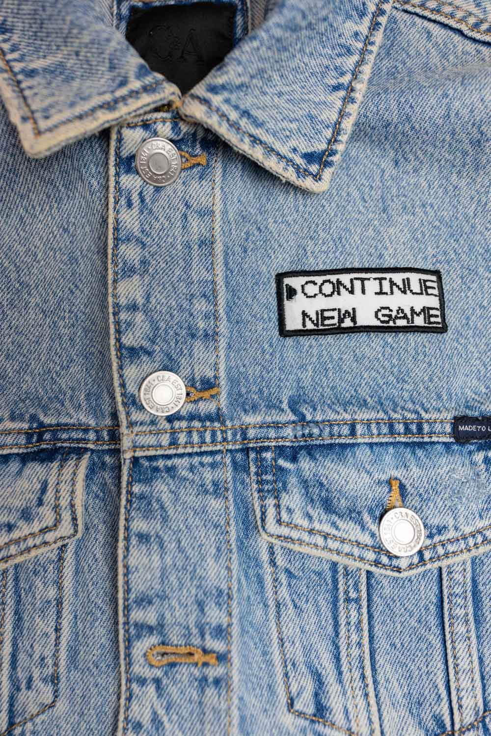 Patch brodat negru cu mesajul „Continue New Game”, un accesoriu inspirat de jocurile video retro.
