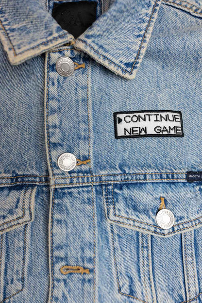 Patch brodat negru cu mesajul „Continue New Game”, un accesoriu inspirat de jocurile video retro.
