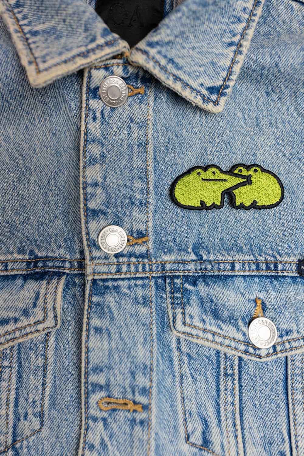 Patch brodat cu design de broască galbenă pe fundal negru, un accesoriu colorat și amuzant pentru haine, genți sau accesorii.