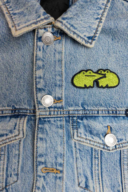 Patch brodat cu design de broască galbenă pe fundal negru, un accesoriu colorat și amuzant pentru haine, genți sau accesorii.