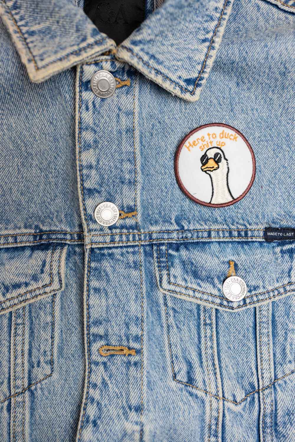Patch brodat rotund cu ilustrație de cap de rață și mesajul amuzant „Here to Duck Shit Up”, un accesoriu ironic și creativ.