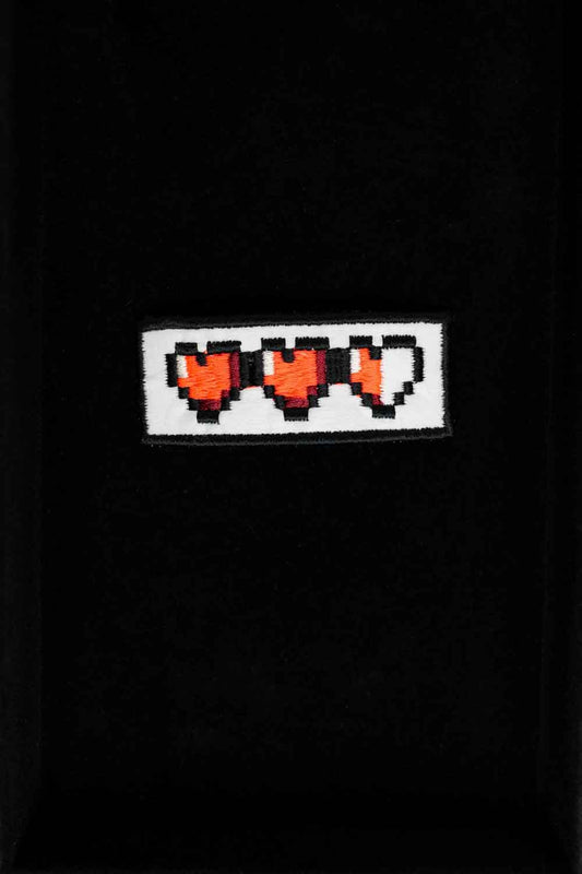 Patch brodat negru cu o serie de inimi pixel roșii, un accesoriu inspirat de stilul gaming retro.