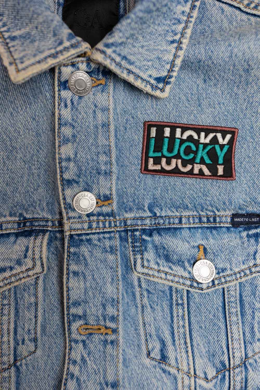 Patch brodat negru cu mesajul „Lucky” repetat într-un font stilizat, un accesoriu modern și expresiv.