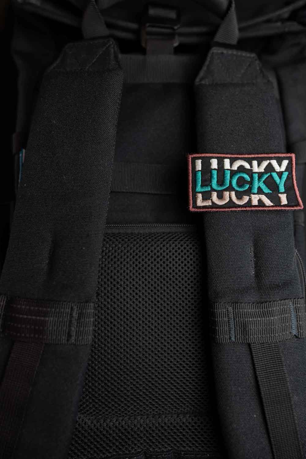 Patch brodat negru cu mesajul „Lucky” repetat într-un font stilizat, un accesoriu modern și expresiv.