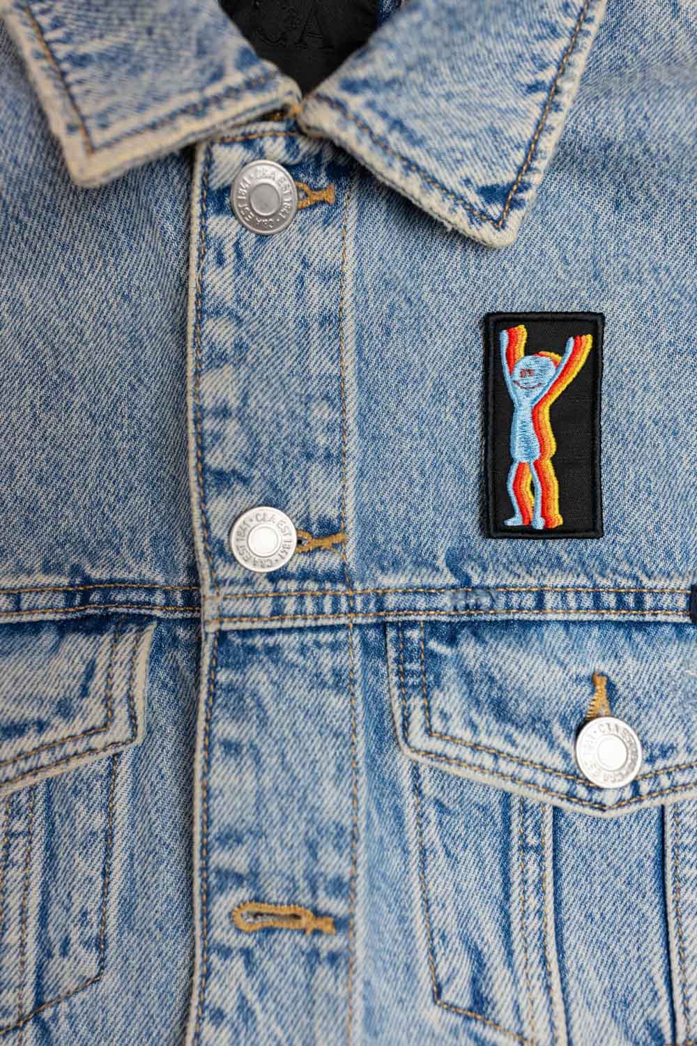 Patch brodat colorat cu un personaj care ține mâinile ridicate, design vesel și amuzant, un accesoriu ideal pentru personalizarea hainelor, genților sau accesoriilor.