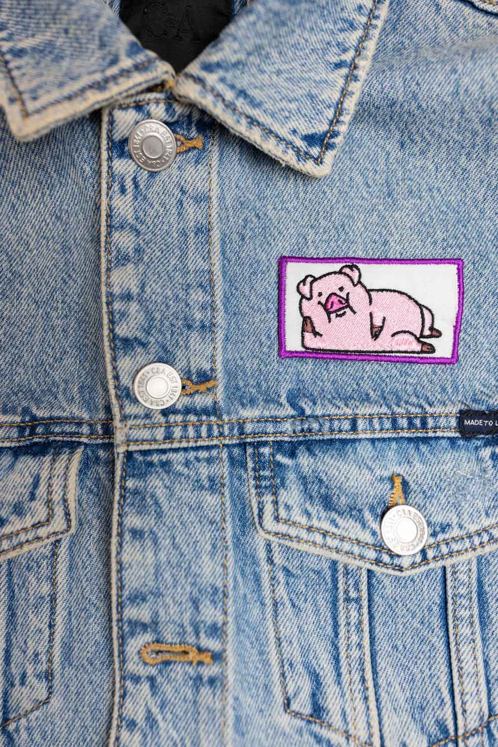 Patch brodat cu purcel roz pe fundal alb și margine mov, un accesoriu simpatic și colorat.