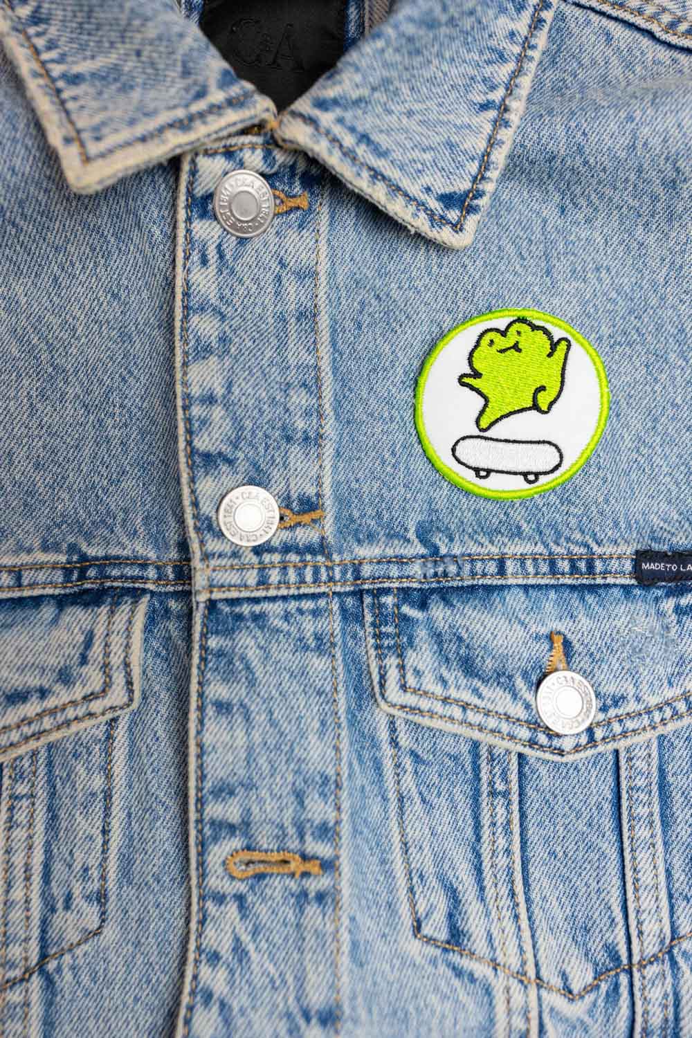 Patch brodat cu personaj stilizat pe skateboard, în culori verde și alb pe fundal negru, un accesoriu pentru iubitorii de skate.