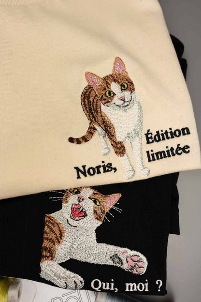 Tricou crem din bumbac organic cu model brodat reprezentând o pisică maro-albă și textul „Noris, Édition limitée”, așezat peste un articol vestimentar negru cu broderie asortată de pisică și textul „Qui, moi ?”.